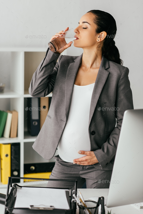 pregnant woman in suit standing in office near table with computer and document tray and drinking