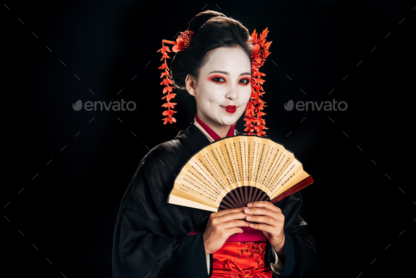 cheerful geisha in black kimono with red flowers in hair holding traditional asian hand fan isolated