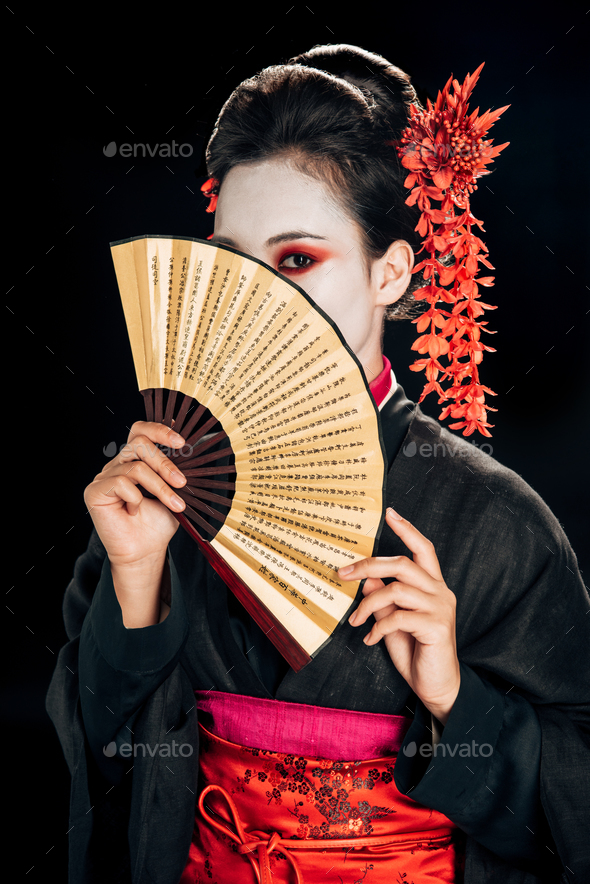 geisha in black kimono with red flowers in hair and obscure face holding traditional asian hand fan