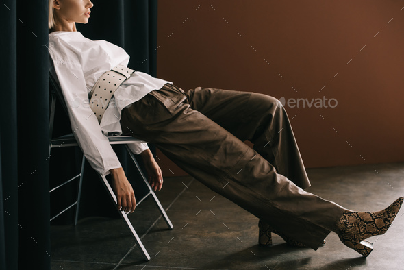 cropped view of stylish woman in white blouse and boots with snakeskin print sitting on chair near