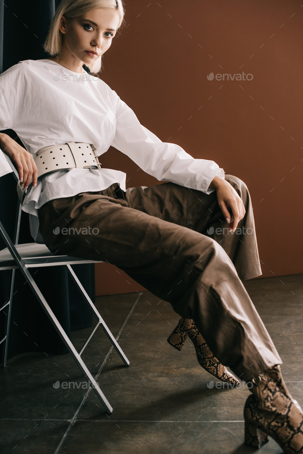 stylish blonde woman in white blouse and boots with snakeskin print sitting on chair near curtain on