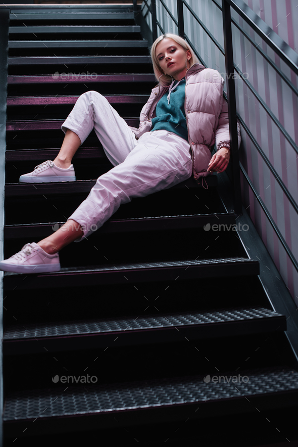 blonde girl in down jacket and sneakers sitting on stairs and looking at Stock Photo by LightFieldStudios