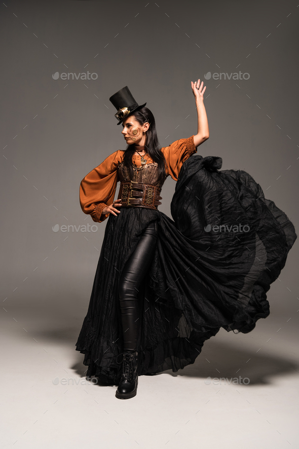 full length view of attractive steampunk woman in top hat with goggles standing with hand on hip on