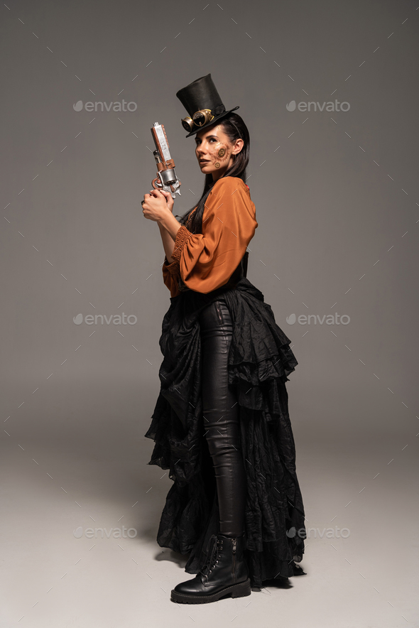 full length view of steampunk woman in top hat with goggles holding pistol on grey