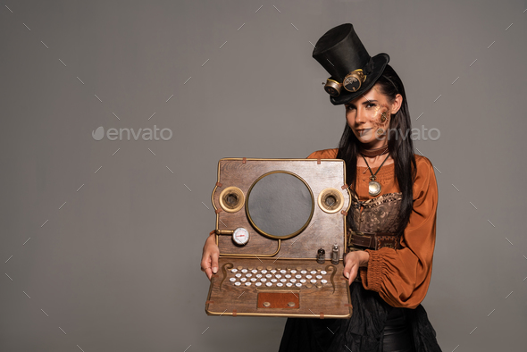smiling woman in top hat with goggles showing steampunk laptop isolated on grey