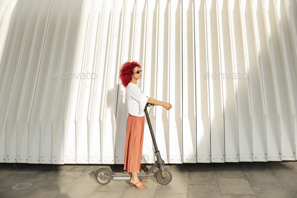 an elegant woman with red afro hair rests leaning on an electric skateboard