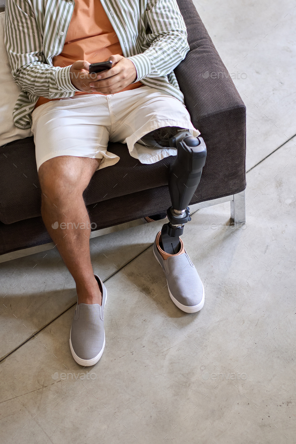 Amputee man with leg prosthesis sitting on sofa at home using phone.