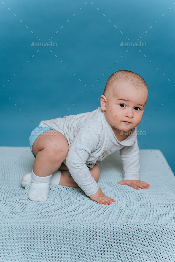 toddler trying to sit looks at camera against blue background. Child boy learning move, stand up