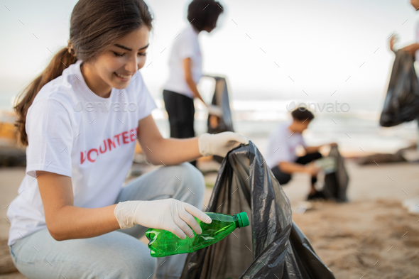 Glad young people volunteers and asian lady in gloves with garbage bag clean up trash on sea beach
