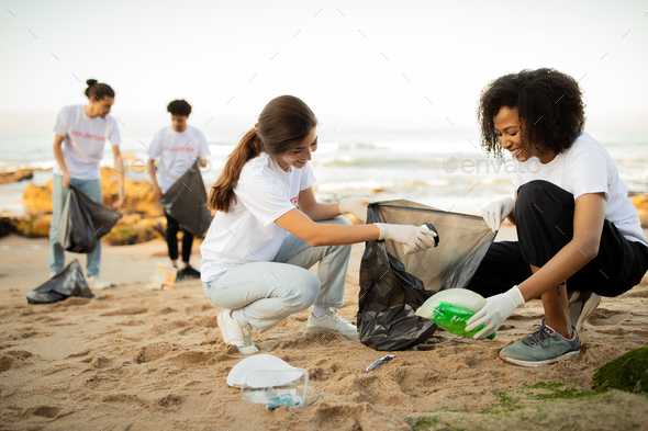 Positive young different people volunteers in gloves with garbage bags clean up trash on sea beach