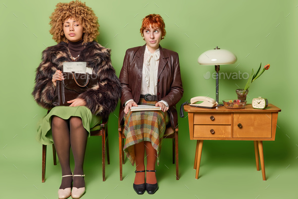 Two women pose on chairs wait for job interview wear fashionable retro clothes pose with papers pose
