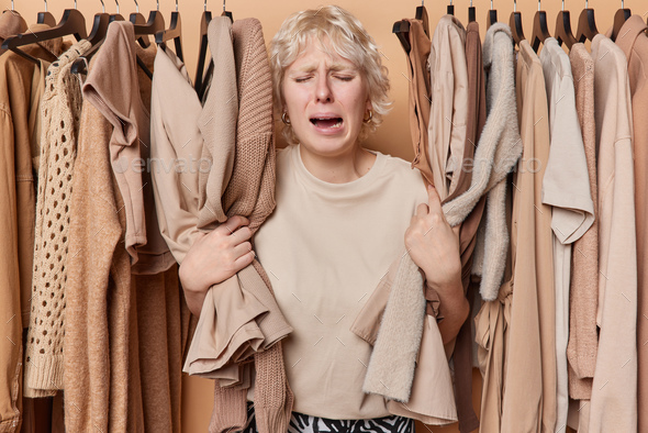 Depressed crying fair haired woman poses among clothing on rack