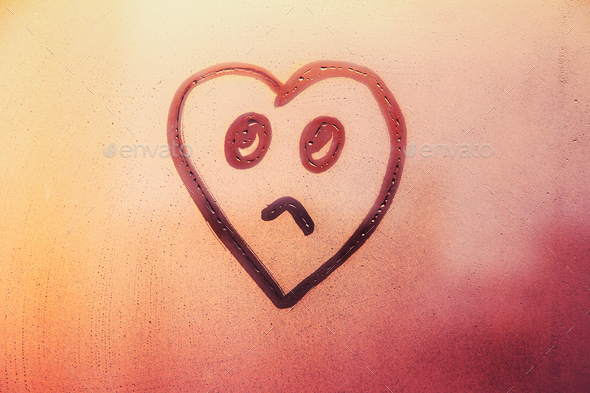 Sad face heart shaped painted on foggy glass window