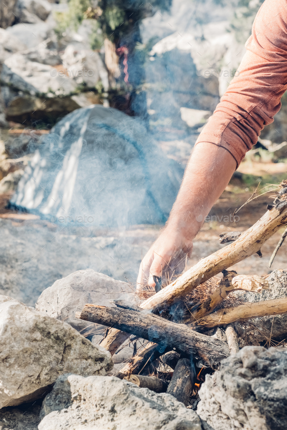 Man hiker putting firewood into flame while making bonfire near tent in mountain - Stock Photo - Images