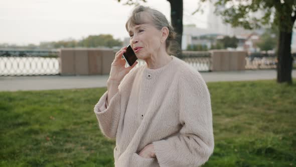 Beautiful Retired Lady Chatting on Cellphone Standing Outdoors in Busy Street in Summer alt