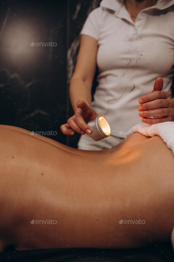Masseur woman drips wax from a candle on the back of the young girl, rubbing massage oil.