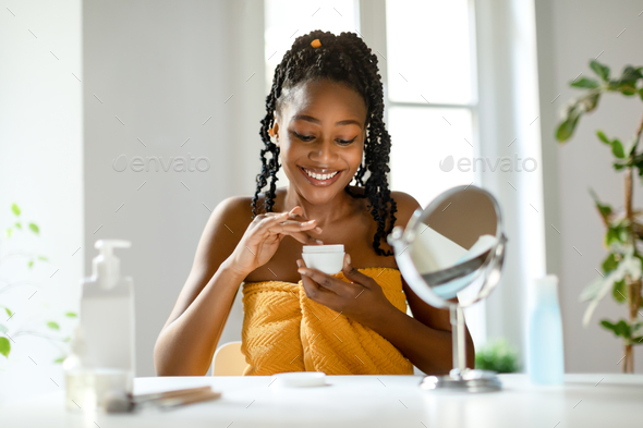Natural beauty routine. Happy black lady holding jar of skin moisturizing cream, sitting at dressing