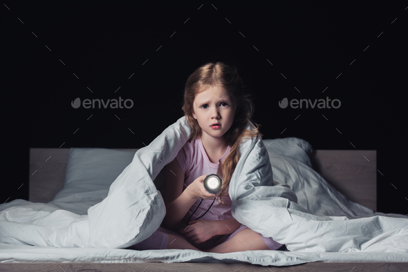scared kid sitting under blanket, holding flashlight and looking at camera isolated on black