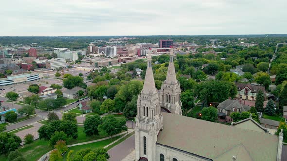 Drone view over church with dual steeples to the city all around with tree lined streets. Cloudy sky