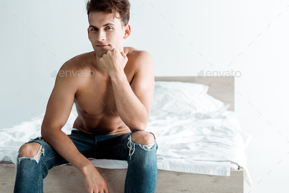 sexy young man in blue jeans sitting on bed at home Stock Photo by