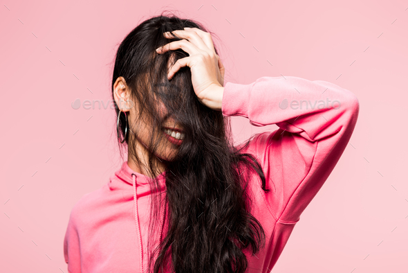 attractive asian woman in pink pullover smiling and obscuring face isolated on pink