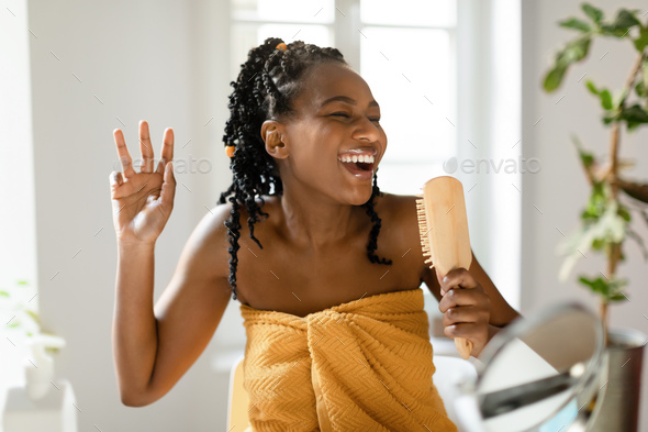 Playful black lady singing while brushing her hair, holding wooden brush as mic, sitting in front of