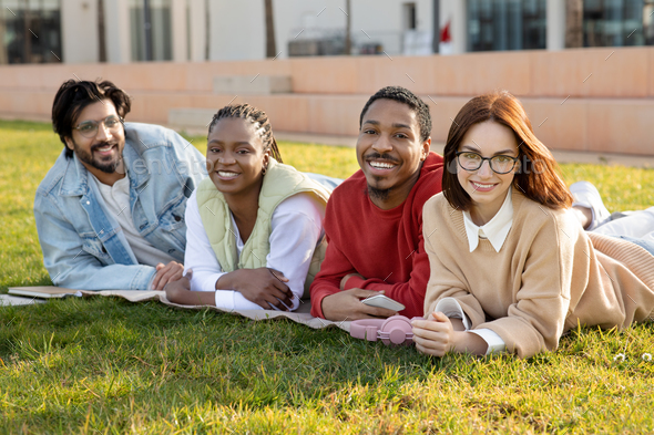 Positive international millennial students exchange, rest outdoor, lie on grass in campus, enjoy