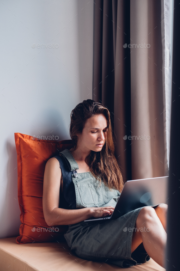 College woman student uses a laptop computer while sitting on a windowsill. - Stock Photo - Images