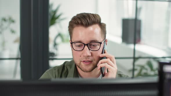 Smiling Male Employee Wearing Glasses Talks to Client on Phone While Working at Computer or alt