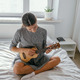 beautiful young woman sitting on the bed and playing ukulele at home - PhotoDune Item for Sale