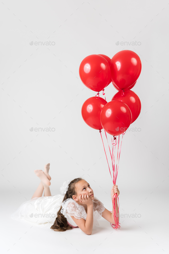 Thoughtful girl ten years old dressed in white dress holding lot of red balloons in hand, looking up