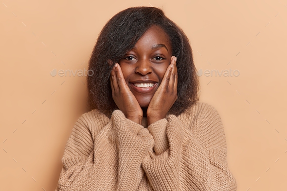 Portrait Of Cheerful Dark Skinned African Woman Keeps Hands On Cheeks Smiles Broadly Glad To