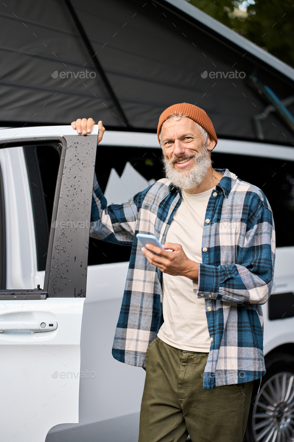 Happy elder man standing near rv camper van on vacation using mobile phone.