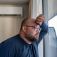 Overweight man hiding at home with working air conditioner in hot weather looking at window. - PhotoDune Item for Sale