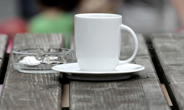 Selective focus shot of a white coffee cup on a plate with an ash pot on the side on a wooden table