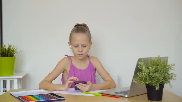 Serious Schoolgirl Sitting at Table with Laptop and Textbook and Doing Homework