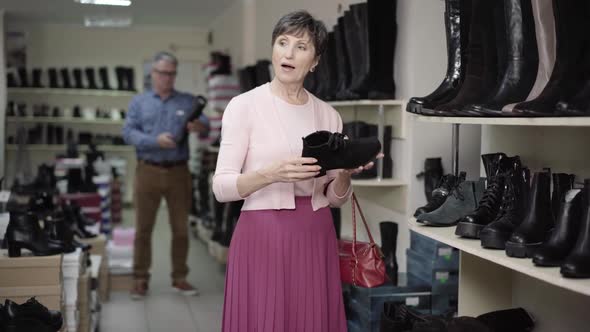 Beautiful Senior Woman Choosing Shoes in Store and Calling Foe Shop Assistant As Blurred Man alt