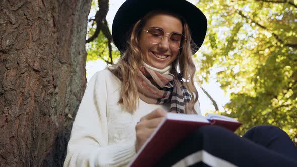 Cute Blonde Woman in the Hat Sitting Near Tree in the Park alt