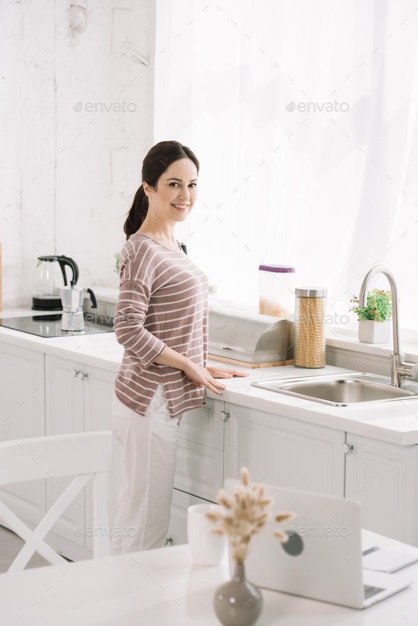 selective focus of kitchen table with laptop near smiling woman standing near sink