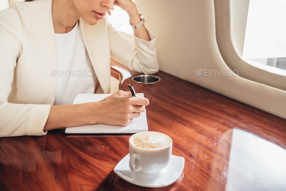 cropped view of businesswoman in suit writing in notebook in private plane