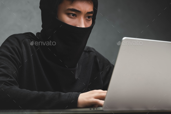 asian hacker with obscured face using laptop and sitting at table