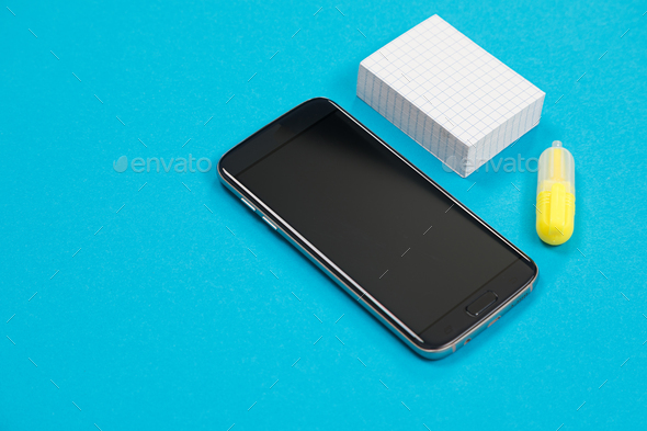 Black smartphone, a pile of scratch paper and one yellow textliner on blue background isolated