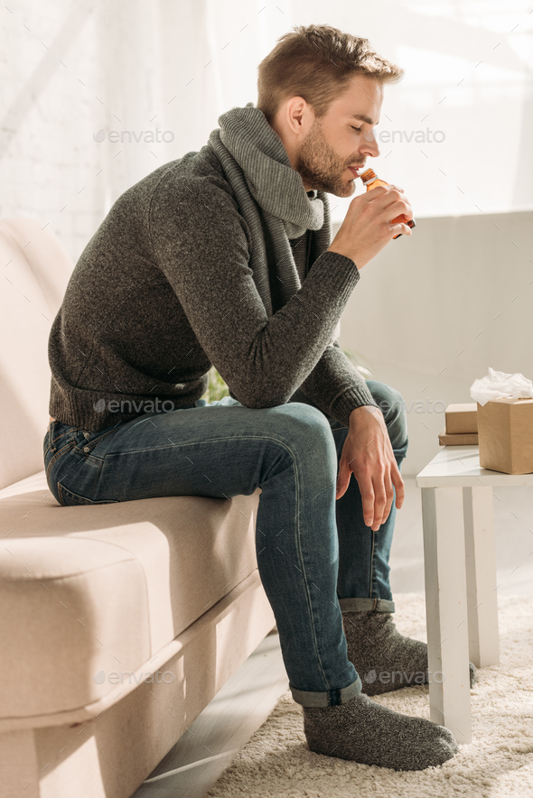 sick man in warm scarf and socks sitting on sofa and drinking cough syrup Stock Photo by