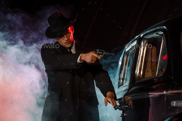 stylish gangster holding gun near retro car on black with smoke