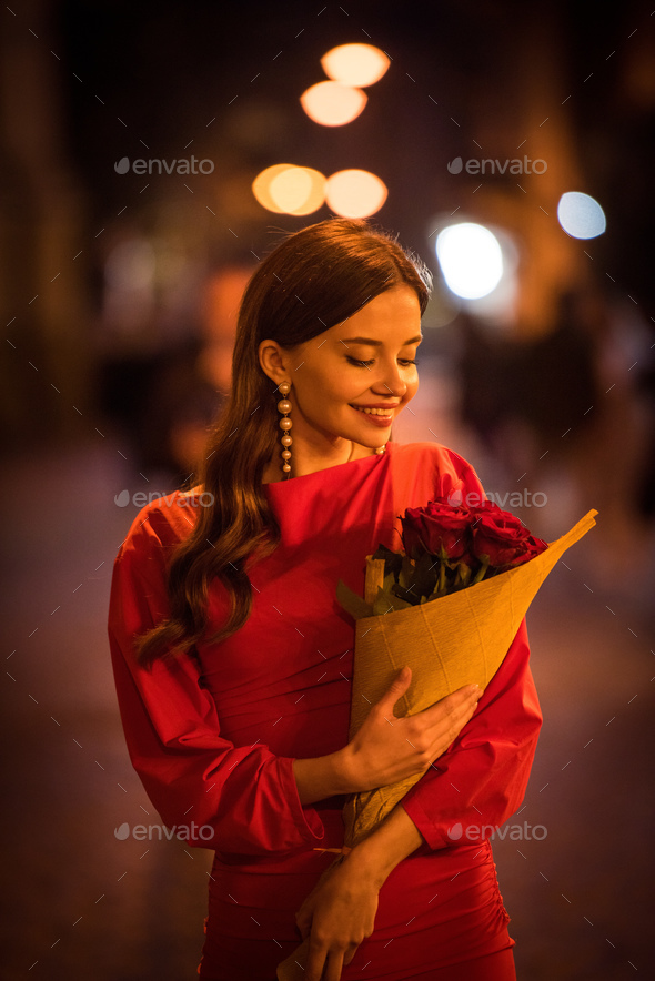 beautiful, smiling girl in pearl earrings holding bouquet of red roses on street at night