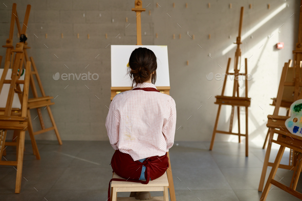 Back view on young woman artist in apron sitting front of blank white canvas