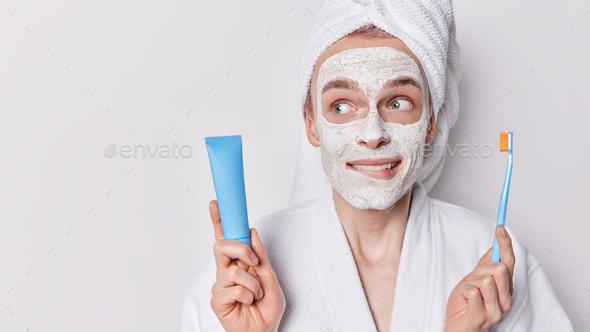 Studio shot of pensive adult man puts on facial mask for reducing fine lines and pores bites lips
