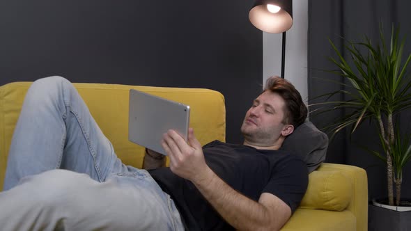 Young Unshaven Handsome Smiling Guy Messaging Online Lying on Sofa with Tablet