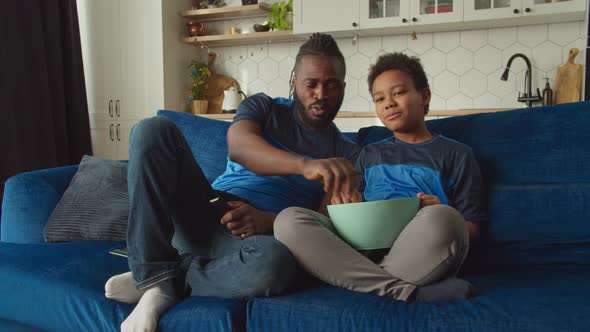Relaxed African American Father and Son Watching Tv and Eating Popcorn alt