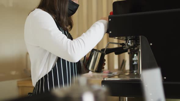 Caucasian waitress wearing face mask standing at coffee machine, preparing froth milk alt